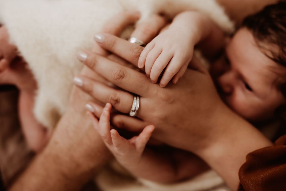 Close-up of tiny baby hand holding parent's fingers with wedding ring visible.