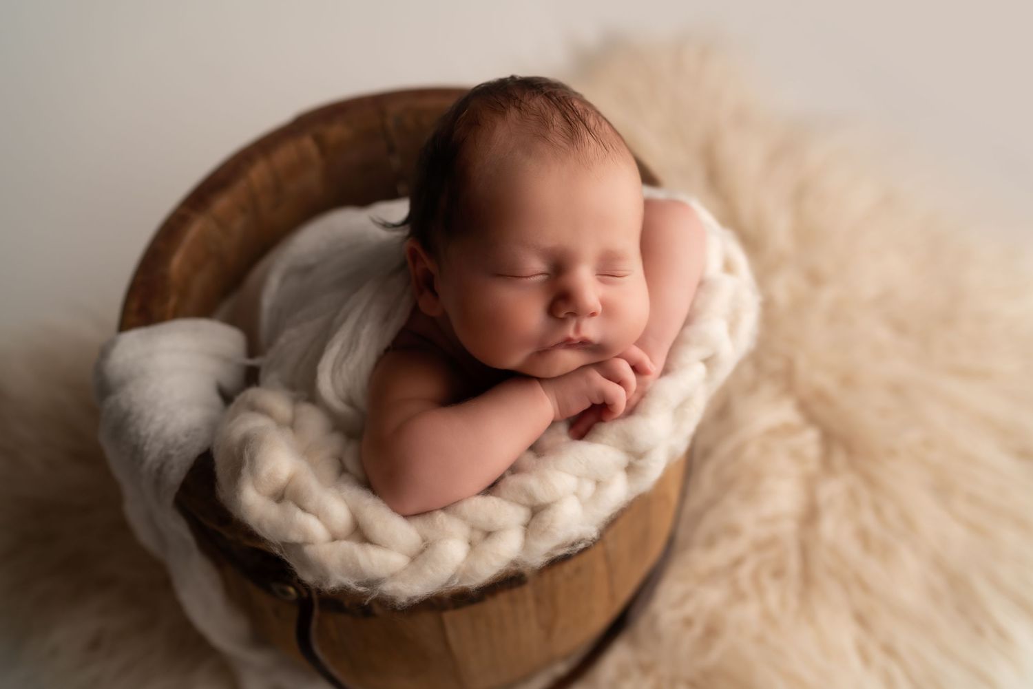 Peaceful newborn sleeping on soft cream-colored blanket in wooden bowl.