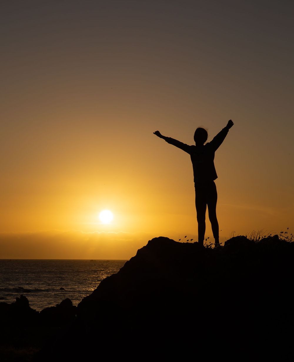 Silhouette of a figure with raised arms against a golden sunset over the ocean.