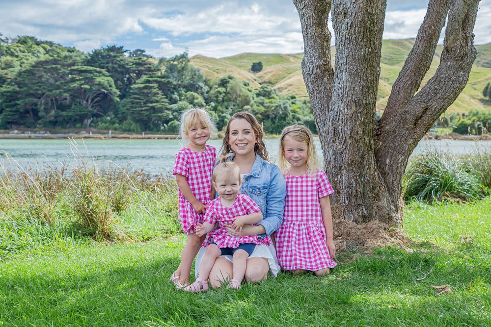 Three girls in pink gingham dresses with woman in denim jacket pose by scenic lake.