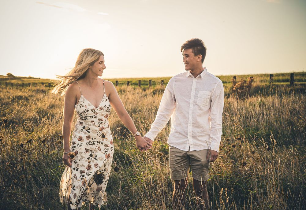 Couple holding hands while walking through tall grass at sunset.