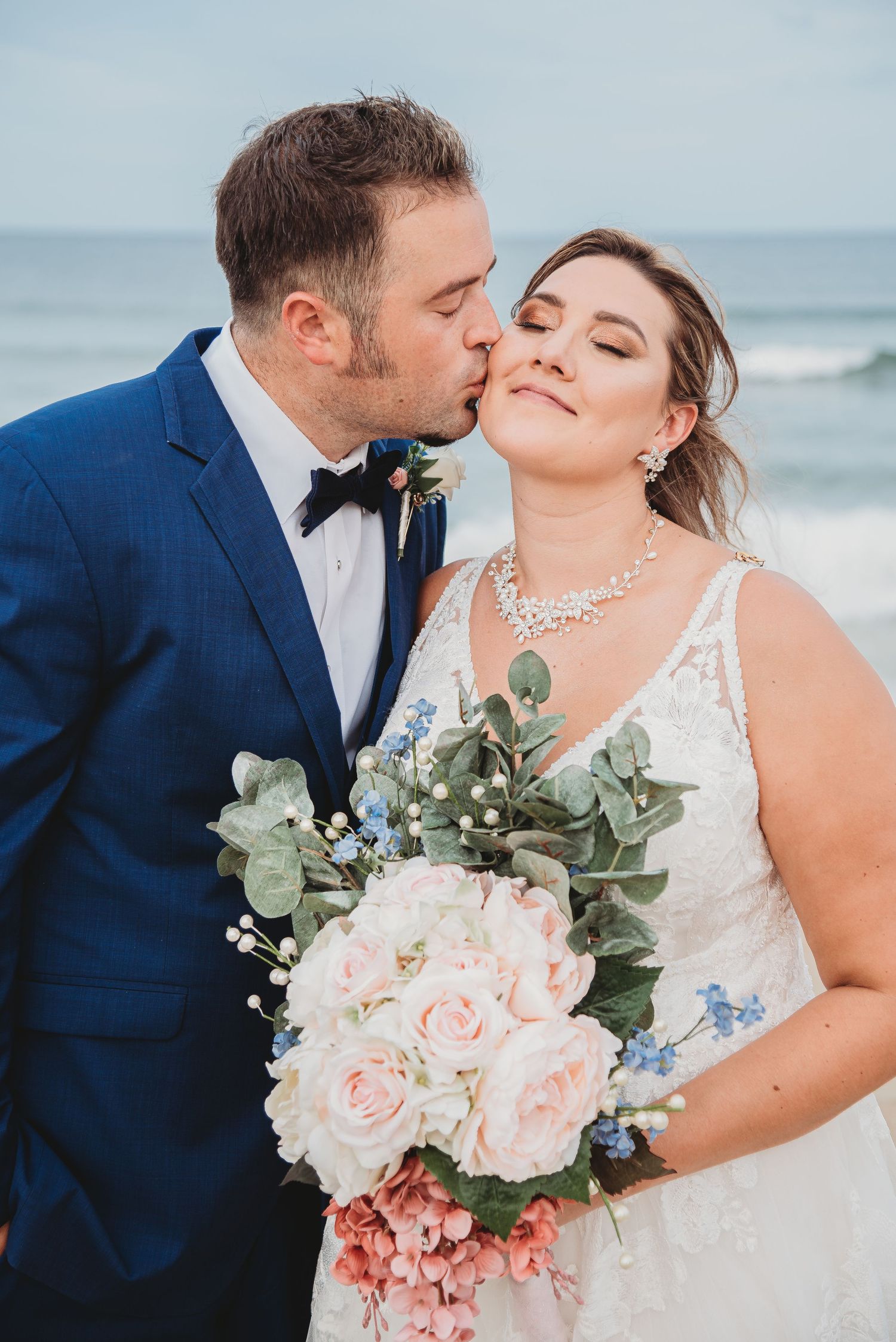 A romantic beach wedding portrait with a bridal bouquet of pink roses and eucalyptus against an ocean backdrop.