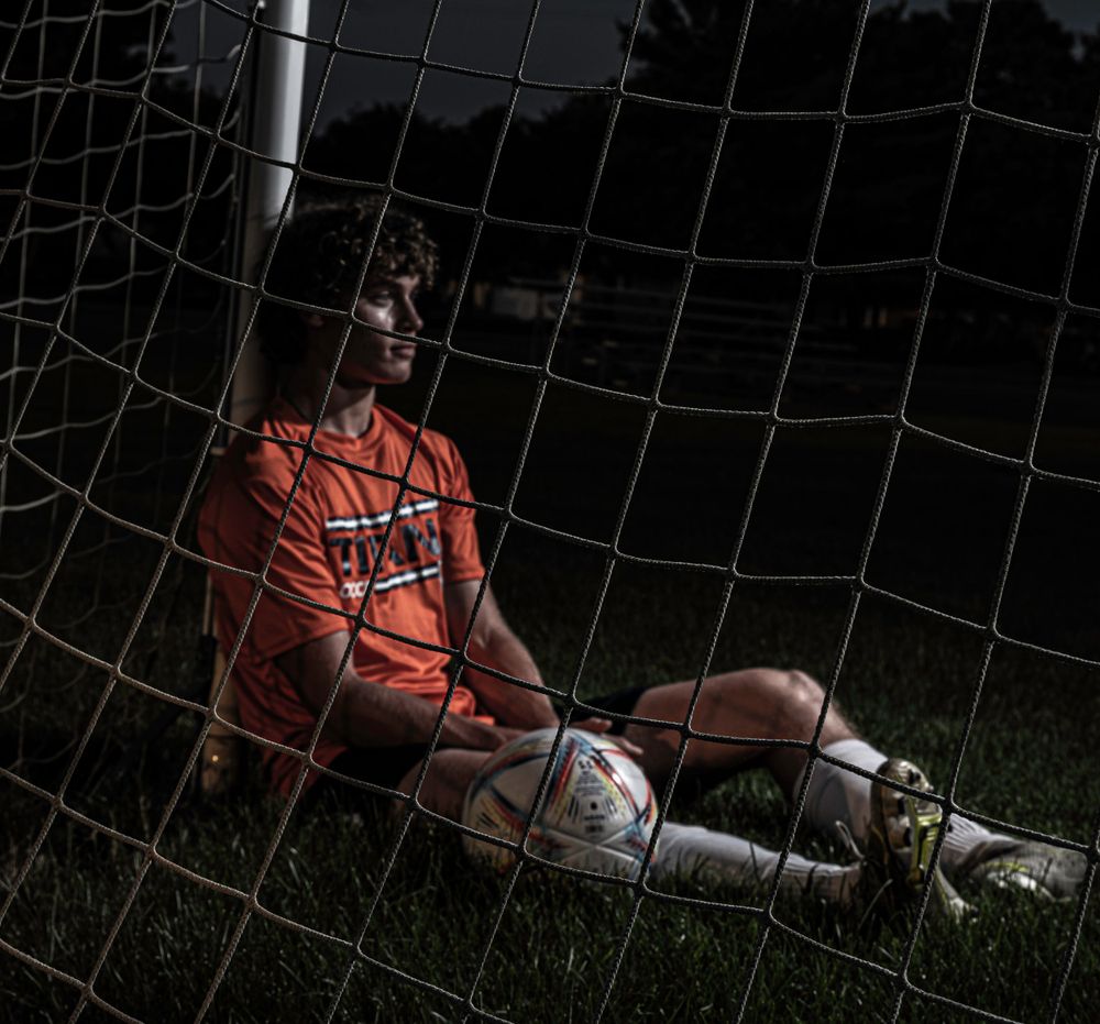 Soccer player in red uniform rests against goal net at dusk in dramatic lighting.
