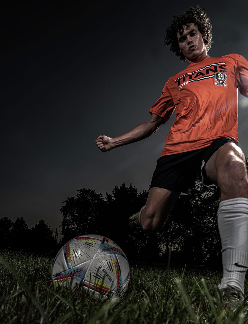 Soccer player in red jersey practices ball control at sunset against dark background.