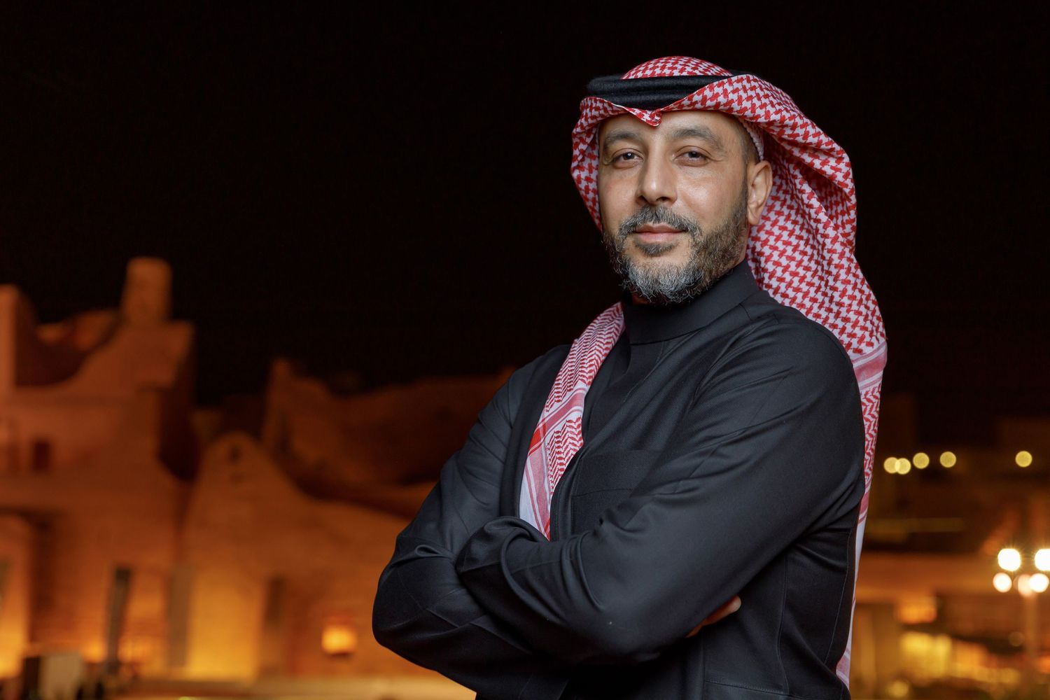 Man in traditional Middle Eastern dress smiles against illuminated building backdrop.