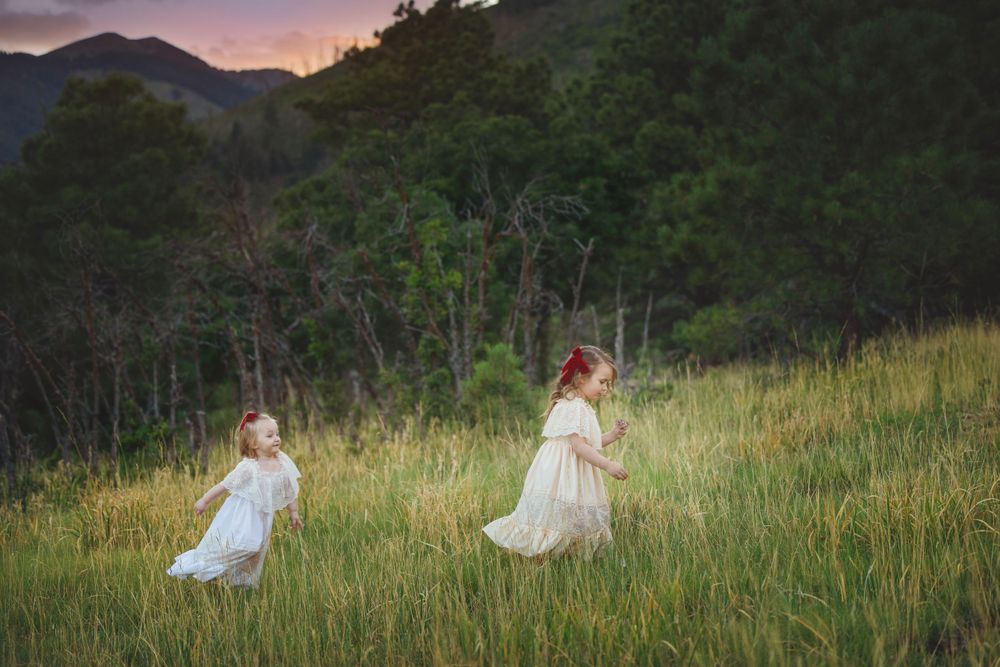 Two young girls in white dresses run through tall grass at sunset with mountains behind.