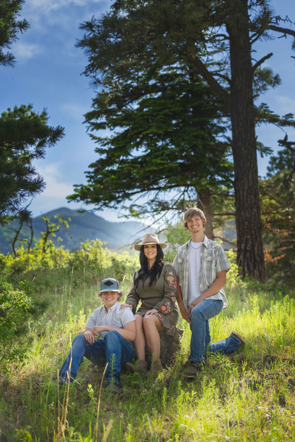 Mother and two sons posing in a sunny mountain forest with tall pines and blue sky behind them.