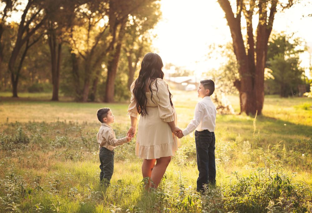 Mother holds hands with two young boys in a sunlit meadow surrounded by golden trees.