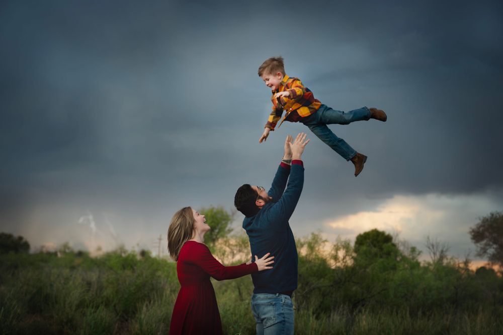 Parents toss smiling child into the air under dramatic stormy skies in an open field.