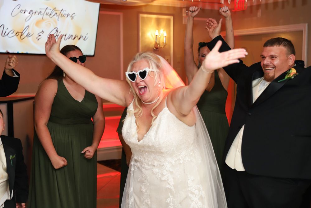 A bride in a white dress and sunglasses dances energetically at her wedding reception.