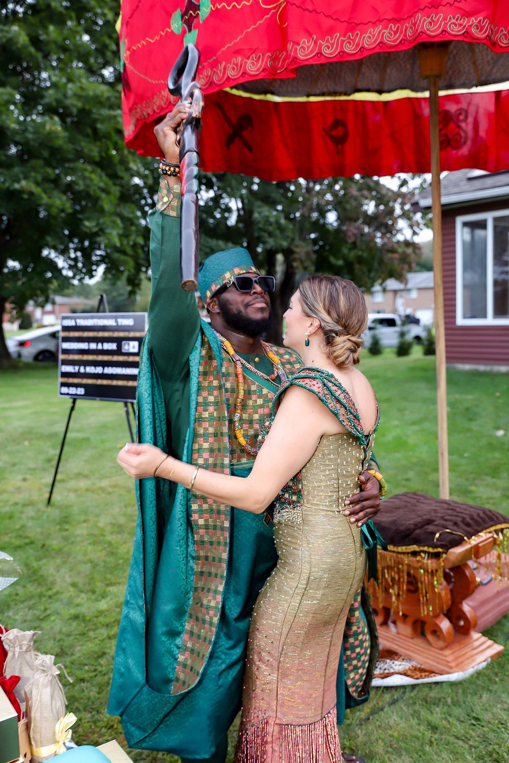 A couple in festive attire celebrates under a red umbrella at an outdoor garden party.