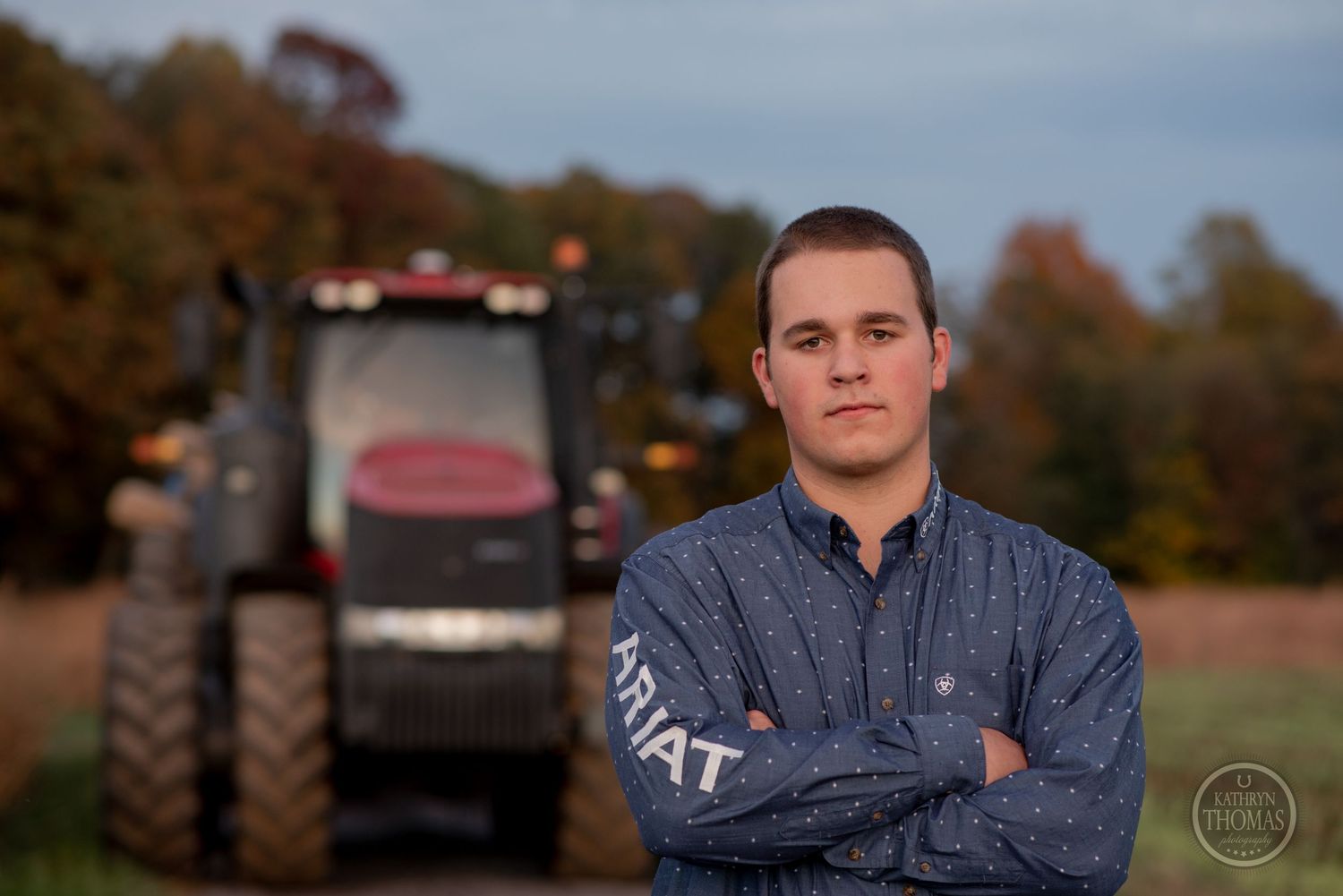 Person in work clothes stands with crossed arms in front of large agricultural tractor.