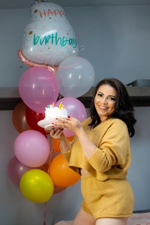Person in yellow sweater celebrating with birthday cake surrounded by colorful balloons.