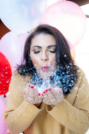 Person in yellow sweater blowing glitter confetti with festive balloons in background.