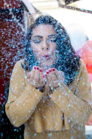 Person in mustard sweater blowing sparkly confetti during celebration photoshoot.