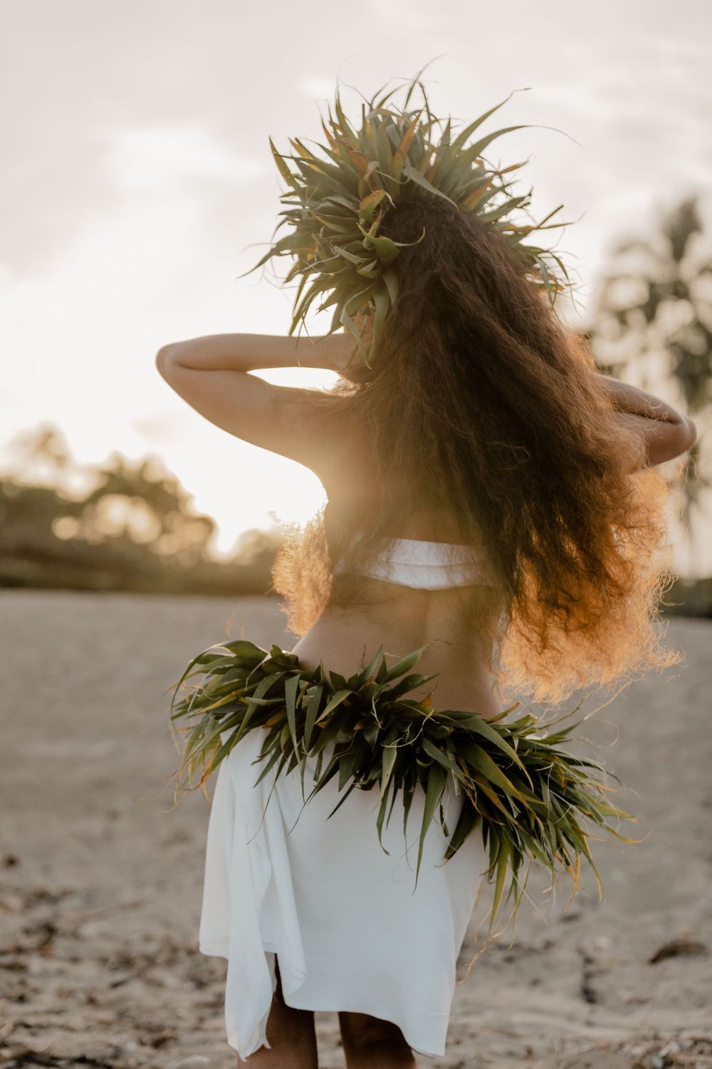 Dancer with flowing hair wearing traditional lei and white dress poses at sunset.