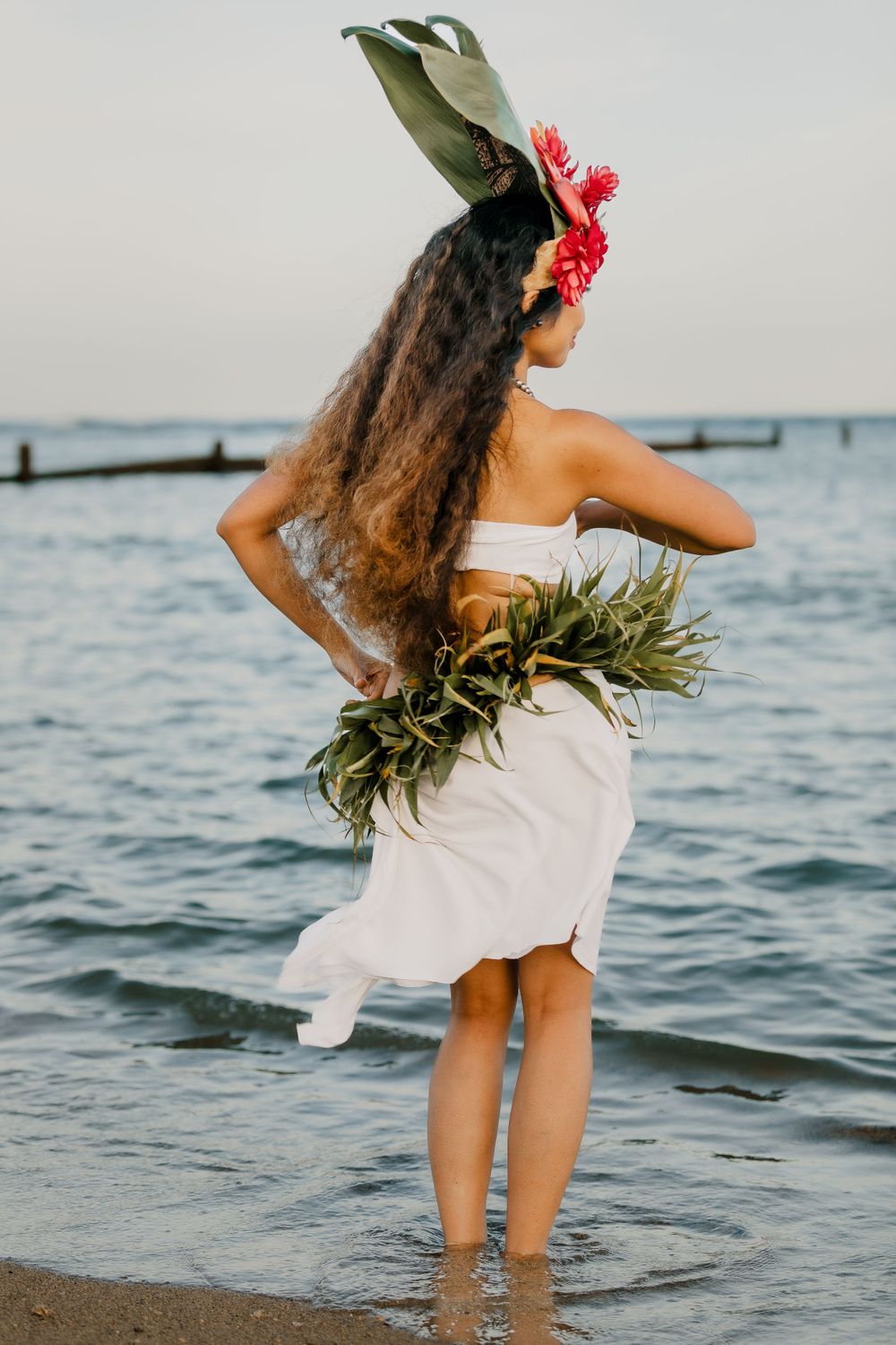 Traditional Hawaiian dancer in white dress and floral headpiece poses by the ocean.