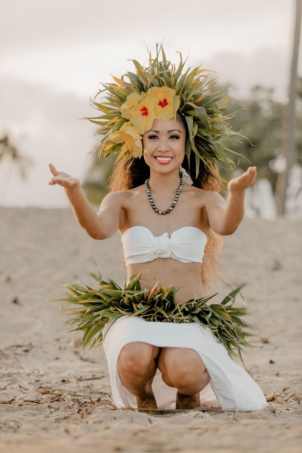 Dancer in traditional Hawaiian attire with elaborate tropical headdress kneels on beach.