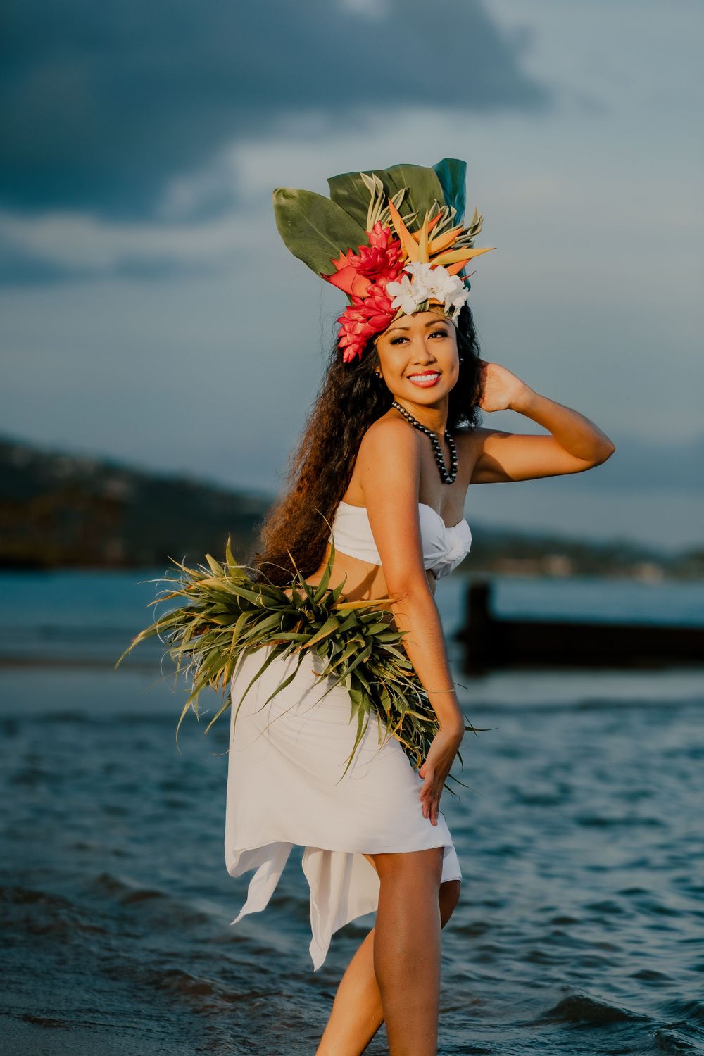 Polynesian dancer in traditional costume poses on beach with tropical floral headpiece.