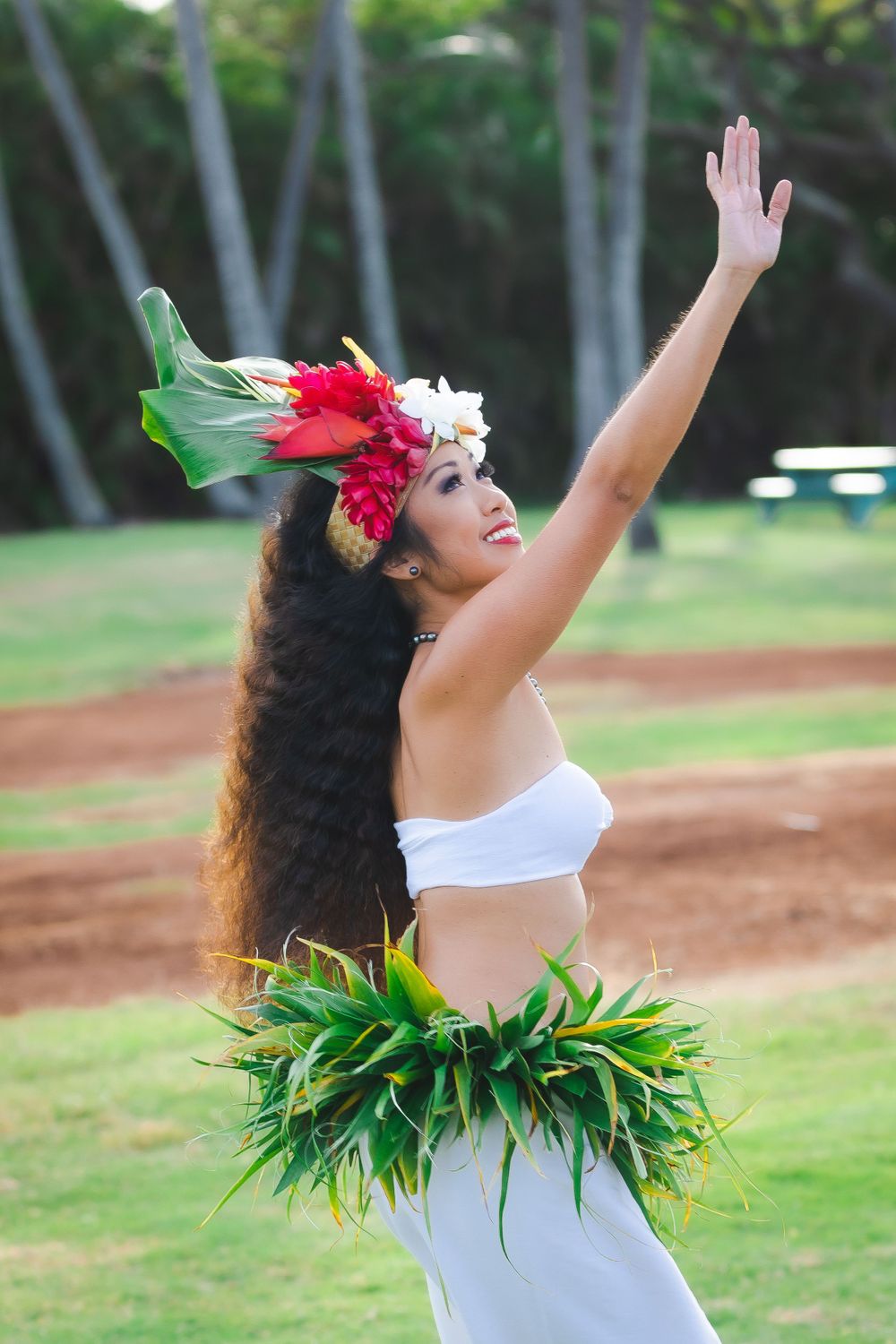 Dancer in traditional Hawaiian attire with arms raised against palm trees.