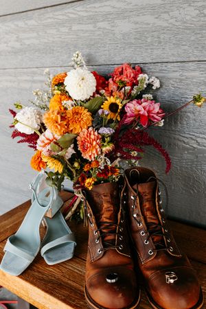 A colorful fall flower arrangement with brown boots and blue heels on wooden surface.