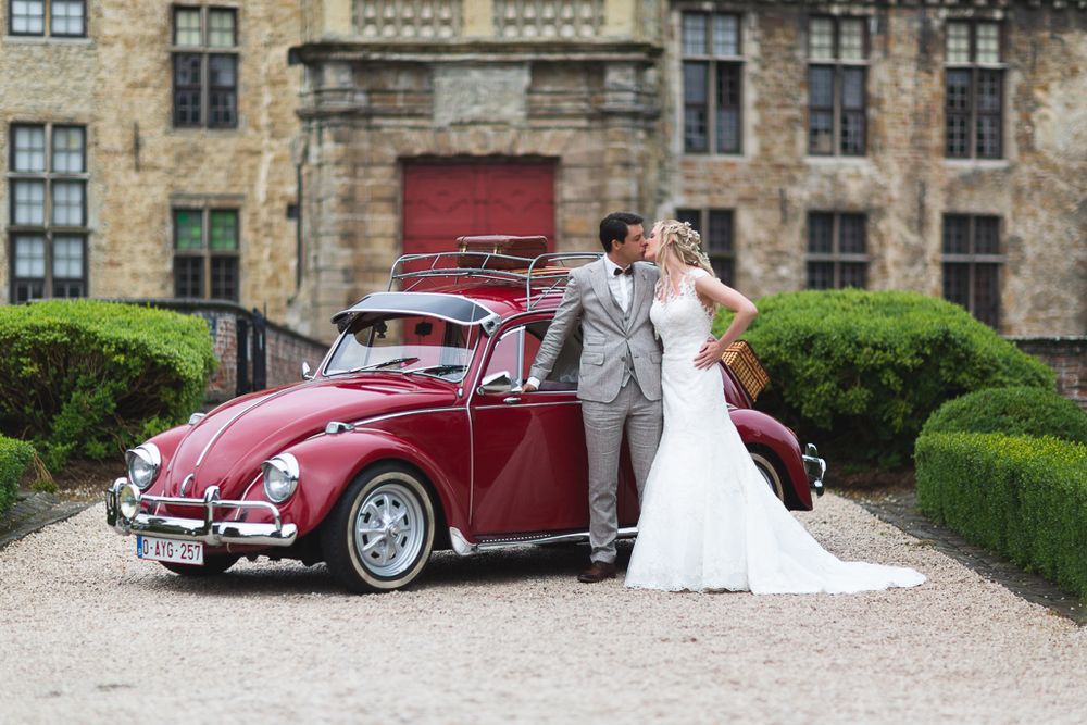Wedding couple poses with classic red Volkswagen Beetle outside historic stone building.