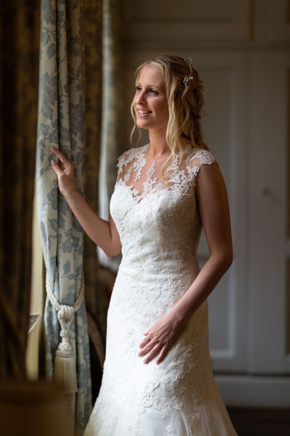 A bride in a lace wedding dress poses elegantly by a window with flowing curtains.