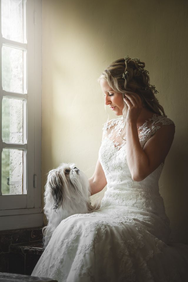 Bride in lace gown sits by window with small white dog in atmospheric lighting.
