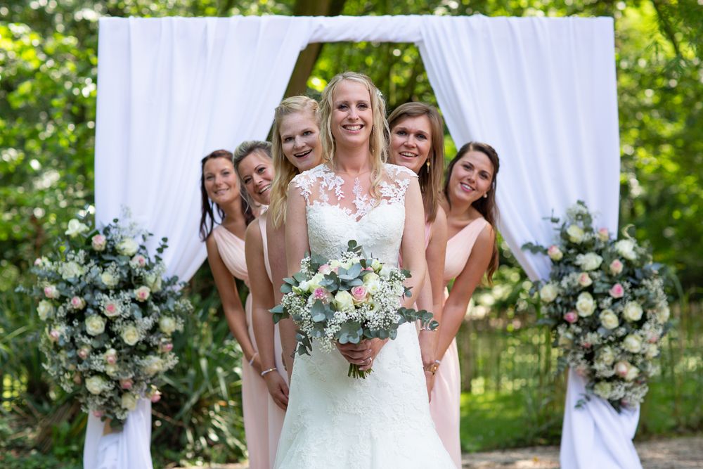 Wedding party poses under white arch decorated with floral arrangements outdoors.