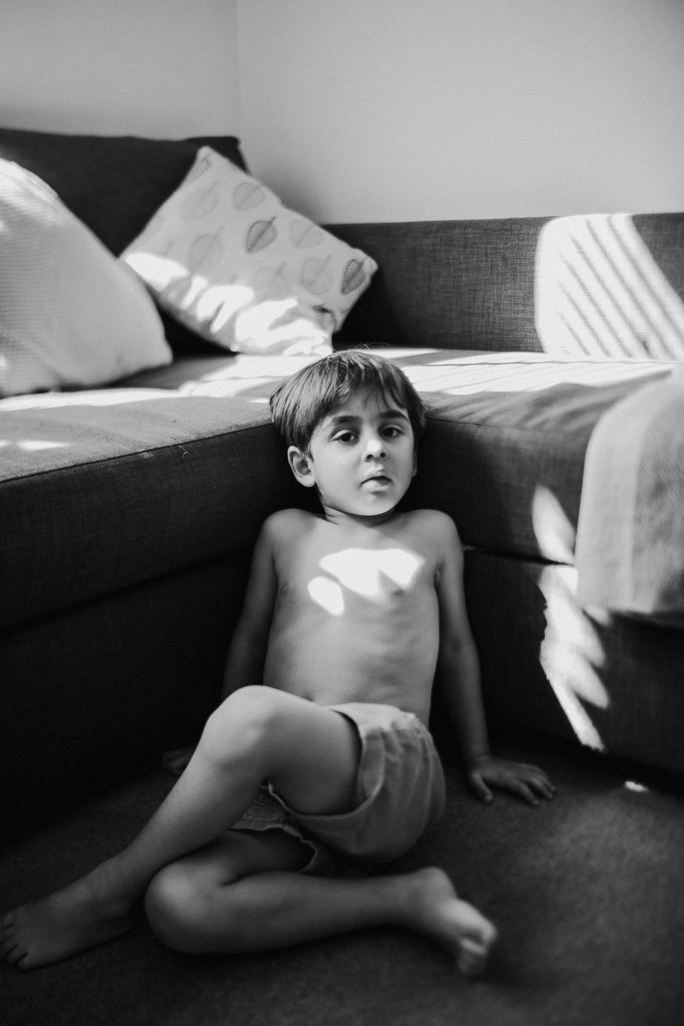 Young shirtless boy sitting on floor leaning against grey sofa in black and white photo with natural light.