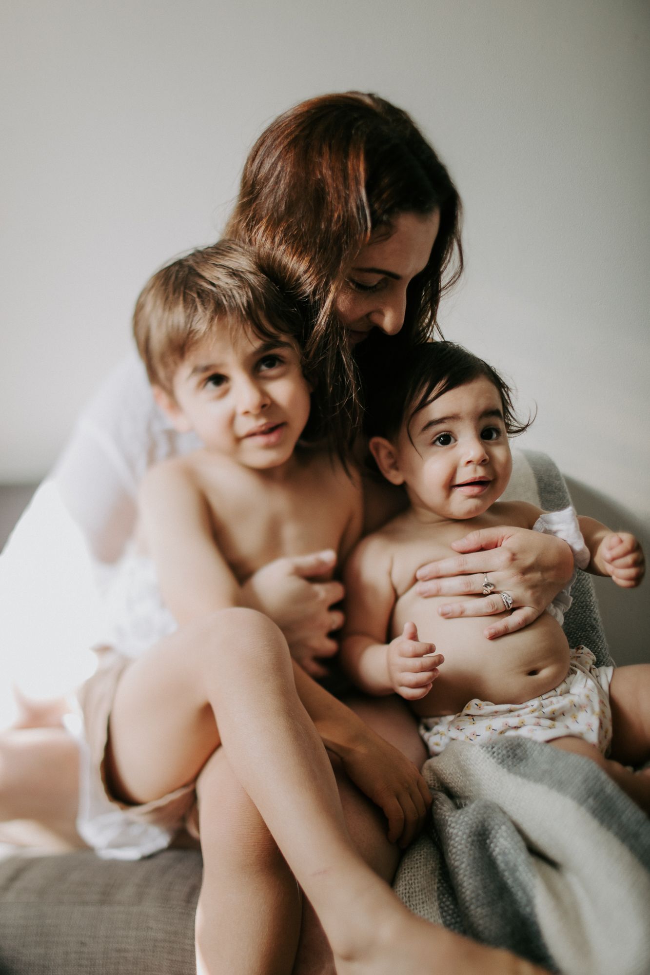 Mother with brown hair embracing two young children on sofa in soft natural light indoor portrait.