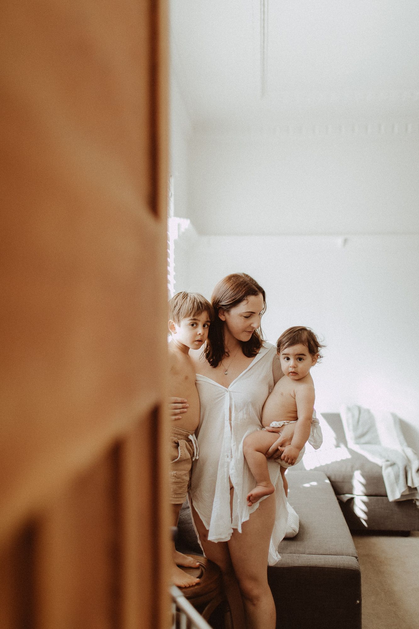 Mother in white dress holding baby with young boy beside her in bright living room.