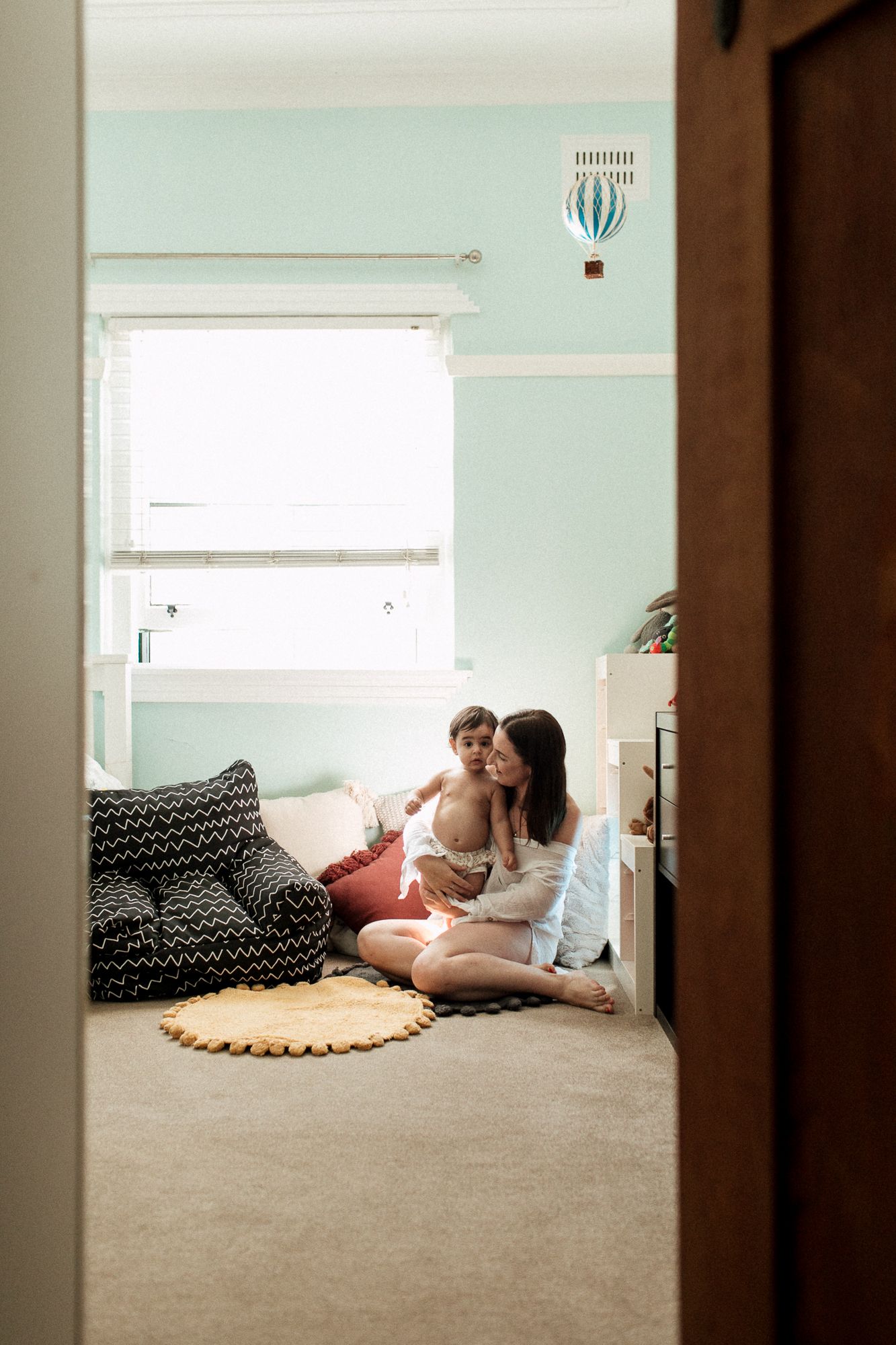 Mother sitting on nursery floor kissing baby in mint green room viewed through doorway.