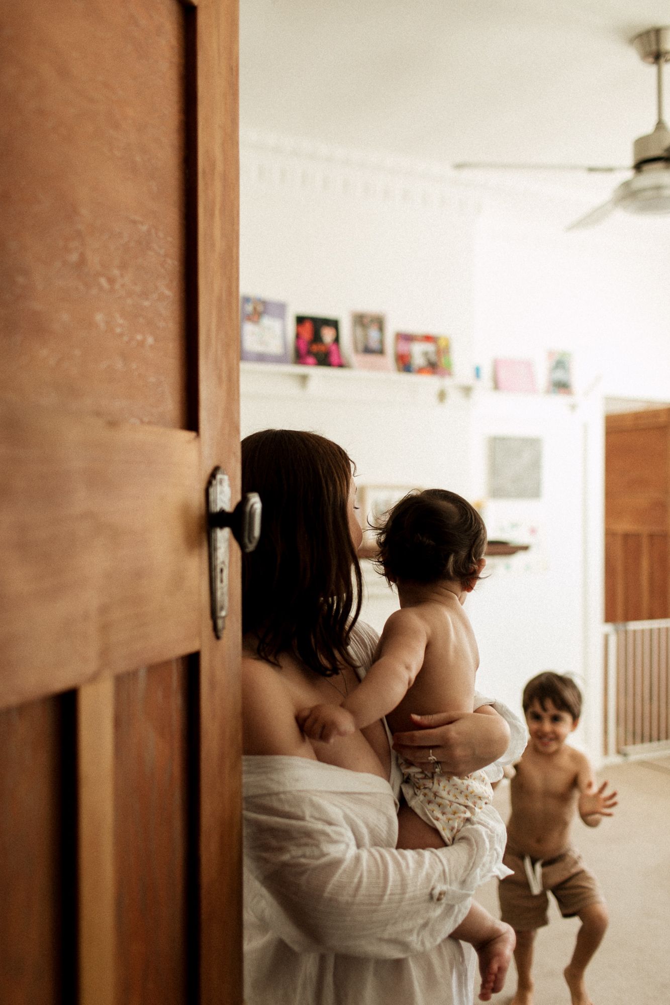 Mother in white robe holding baby while young boy runs in bright home interior through doorway.