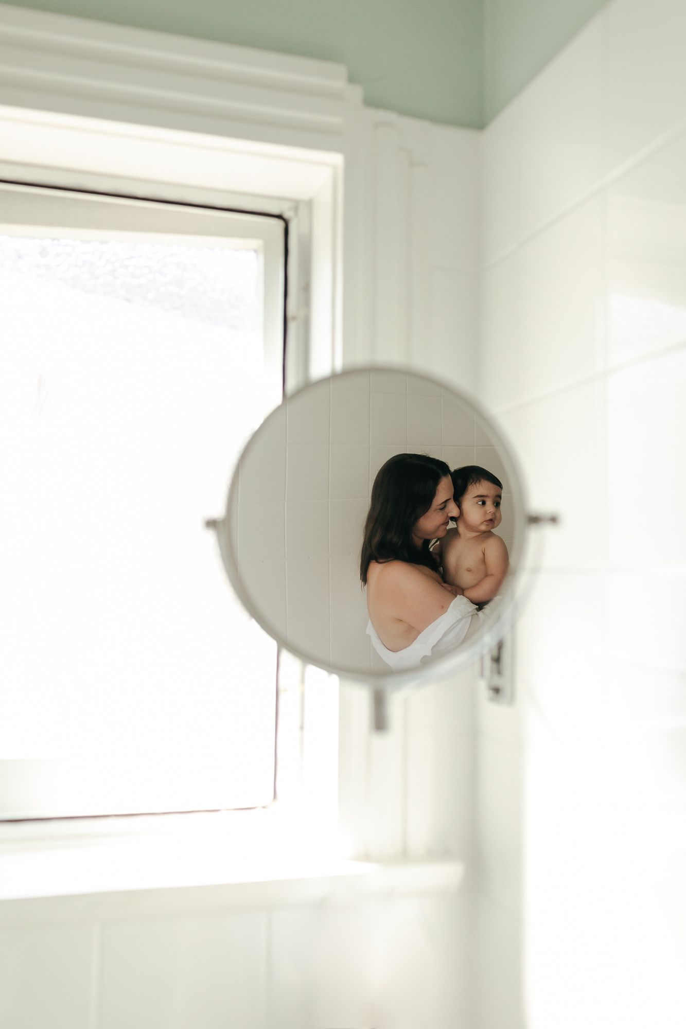 Mother and baby reflected in round bathroom mirror with bright window in white tiled bathroom.