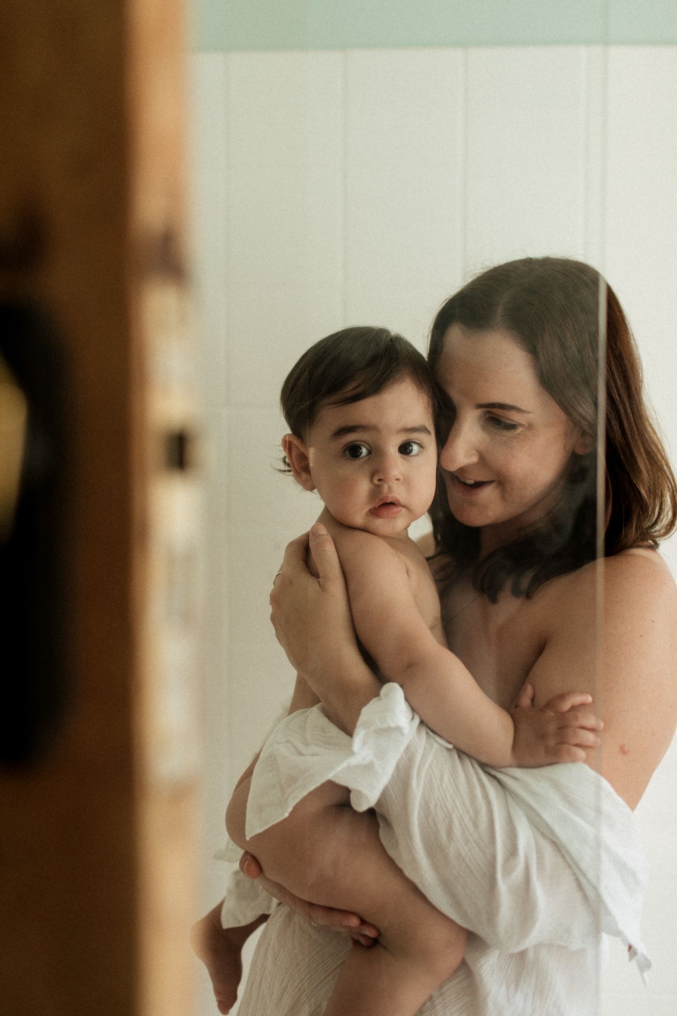 Mother wrapped in white towel holding baby in bathroom mirror reflection with soft natural light.