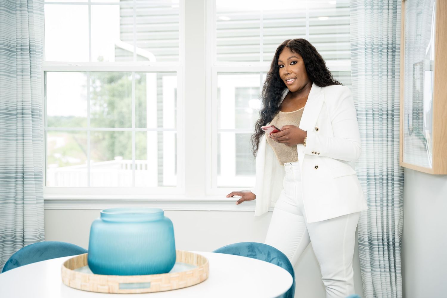 Professional woman in white outfit standing by window with blue vase home decor.