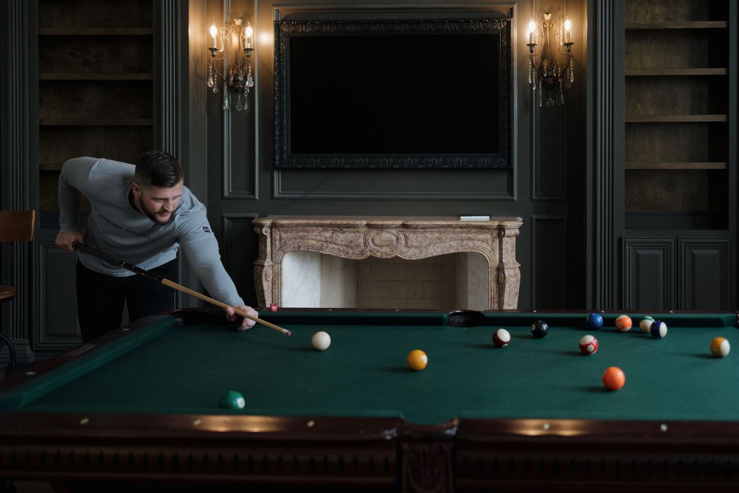 A person plays pool in an elegant room with dark green walls, ornate fireplace, and chandeliers.