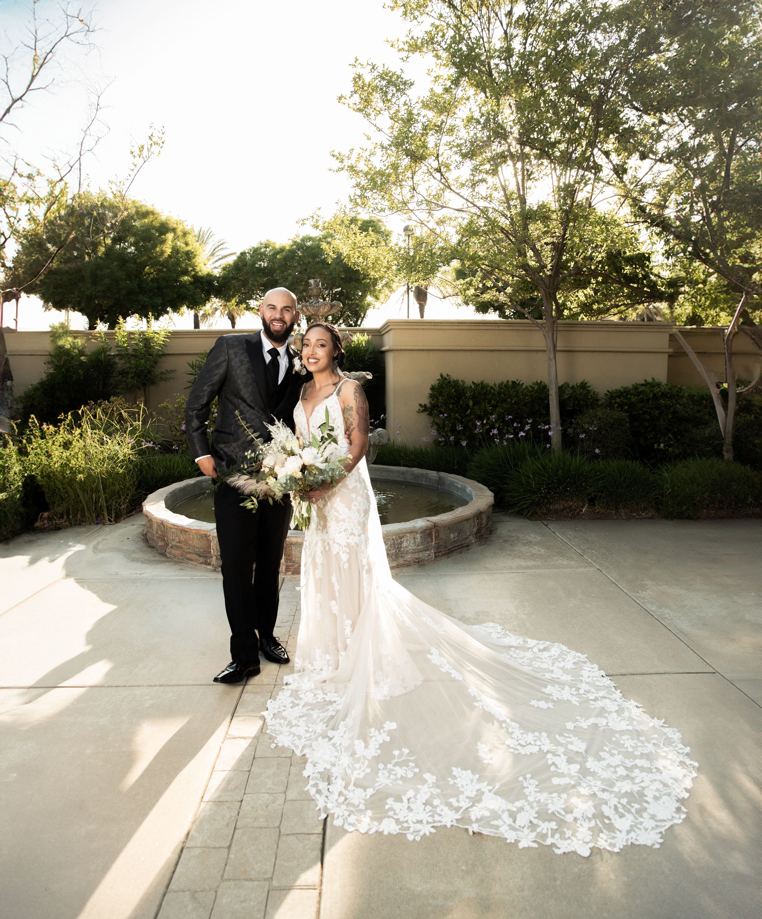 Elegant wedding portrait featuring a bride in lace gown with long train near garden fountain.
