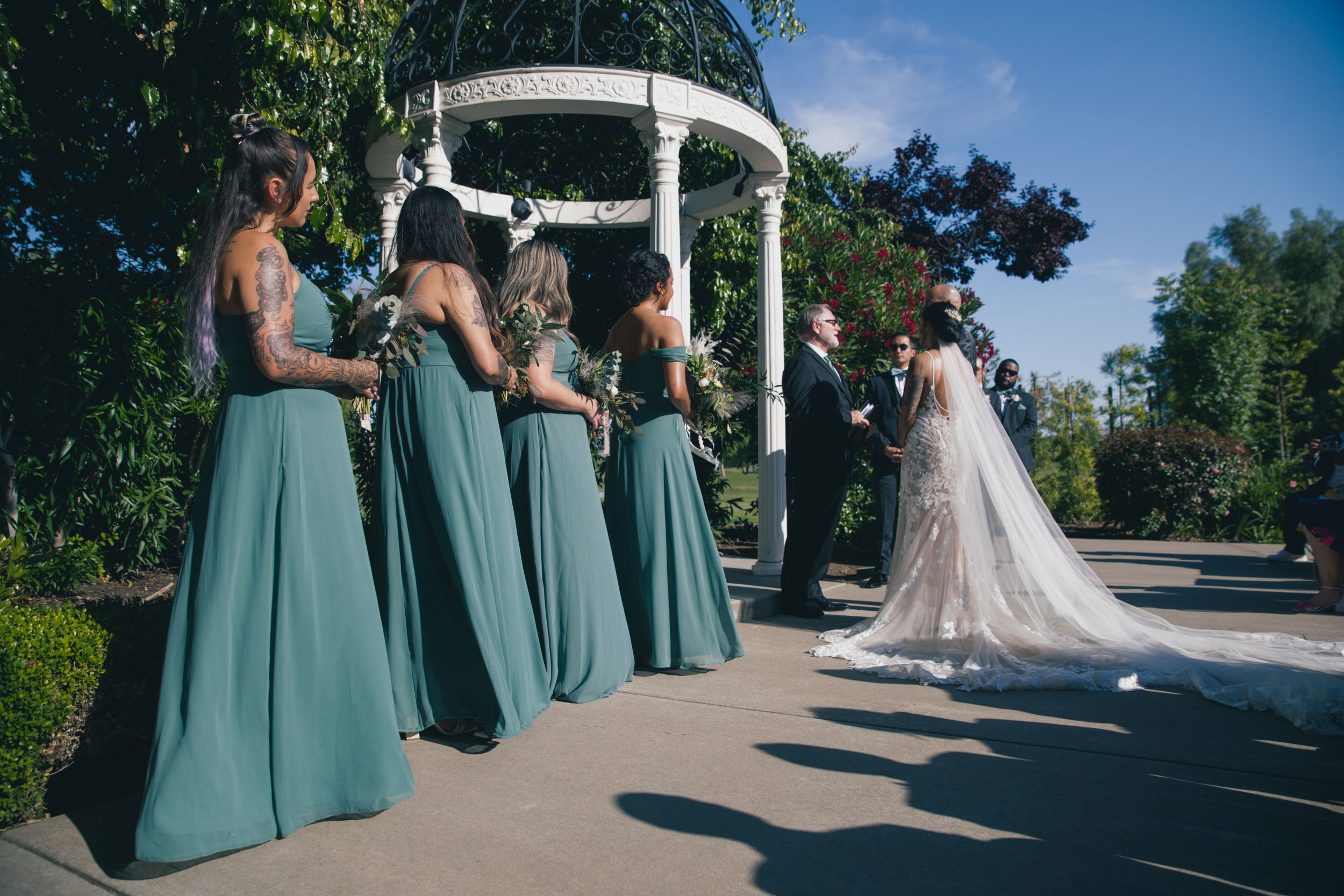 Wedding party lineup with bridesmaids in sage green dresses near white gazebo.