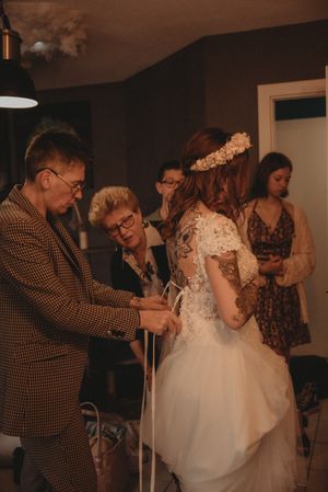 A wedding dress fitting scene with attendees helping adjust the gown in an intimate indoor setting.
