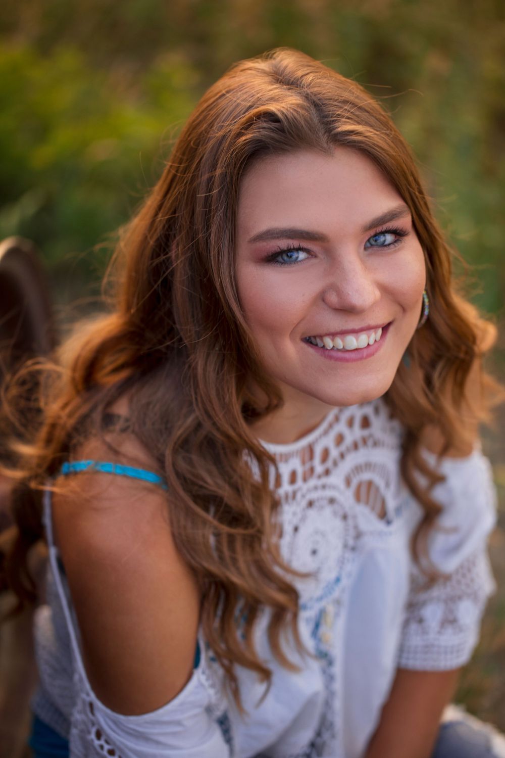 A portrait of a person in a white lace top smiling warmly while outdoors with blurred natural background.