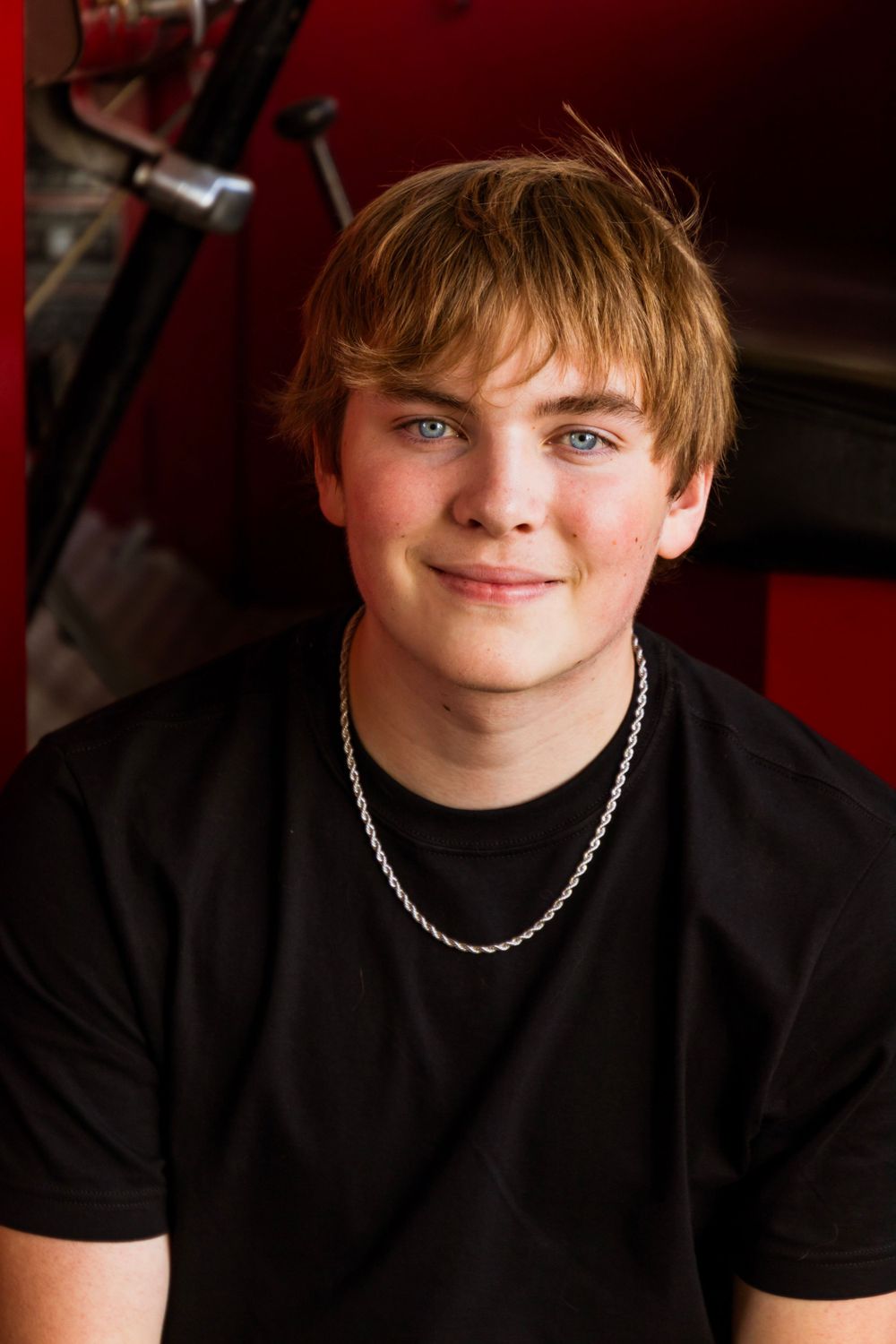 Portrait photo against red background featuring teen wearing black shirt and silver chain necklace.
