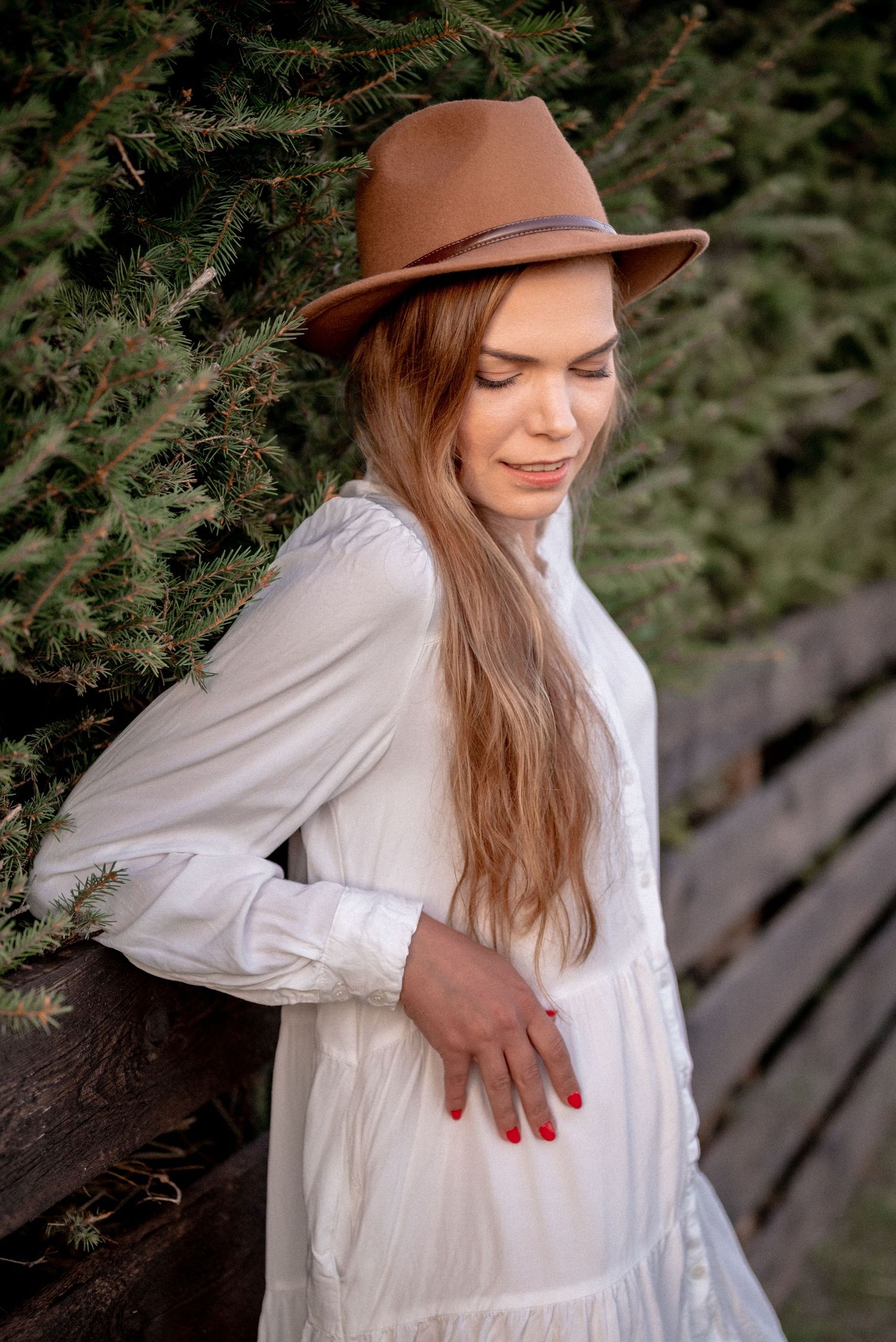 Woman in flowing white dress and brown fedora leans against rustic wooden fence near pine branches.