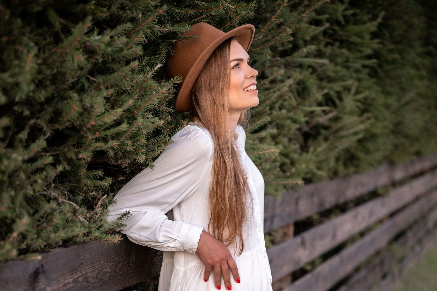Woman in white dress and brown hat gazes upward while leaning on wooden fence outdoors.