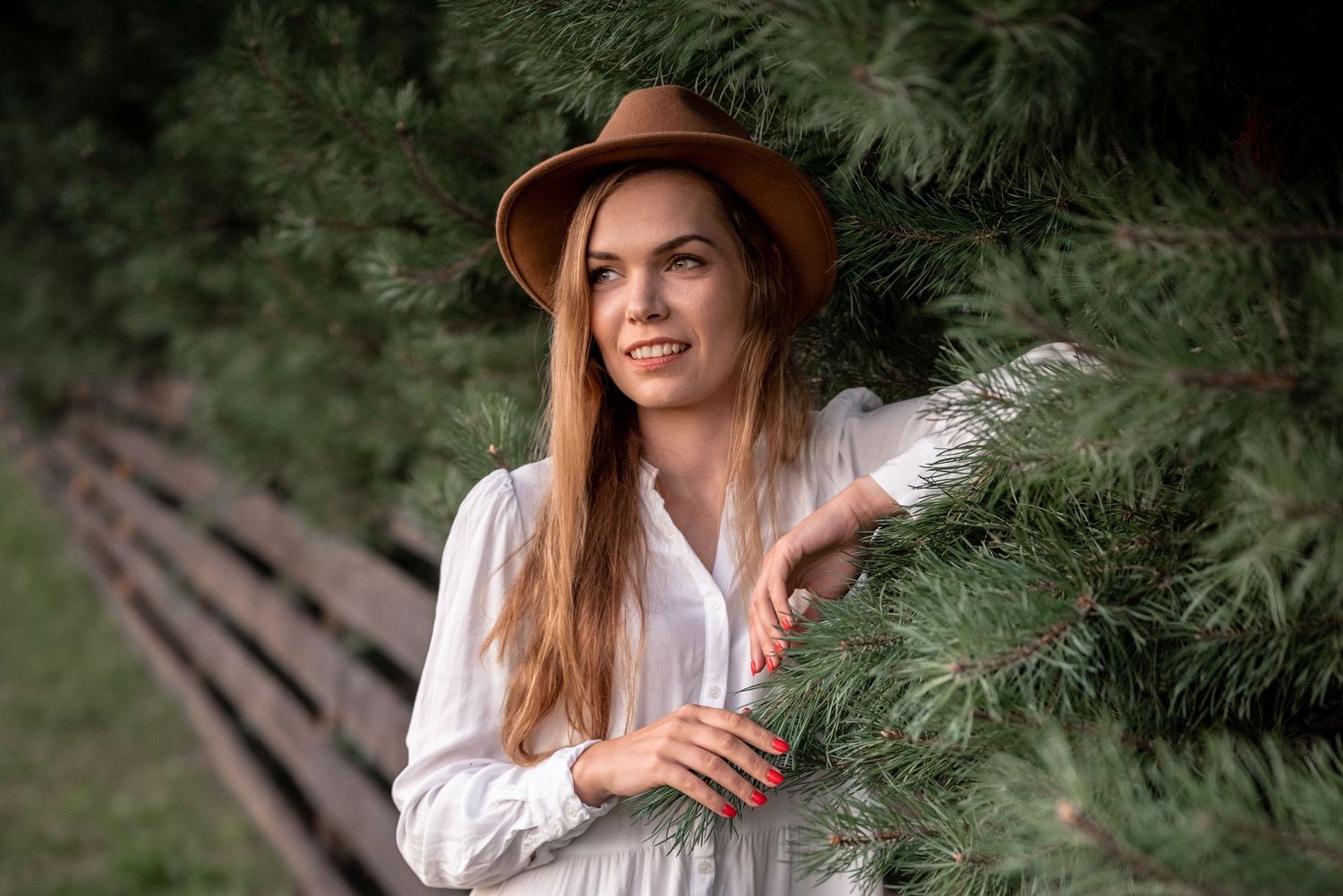 A person in a white blouse and brown hat stands near evergreen trees holding a white feather.