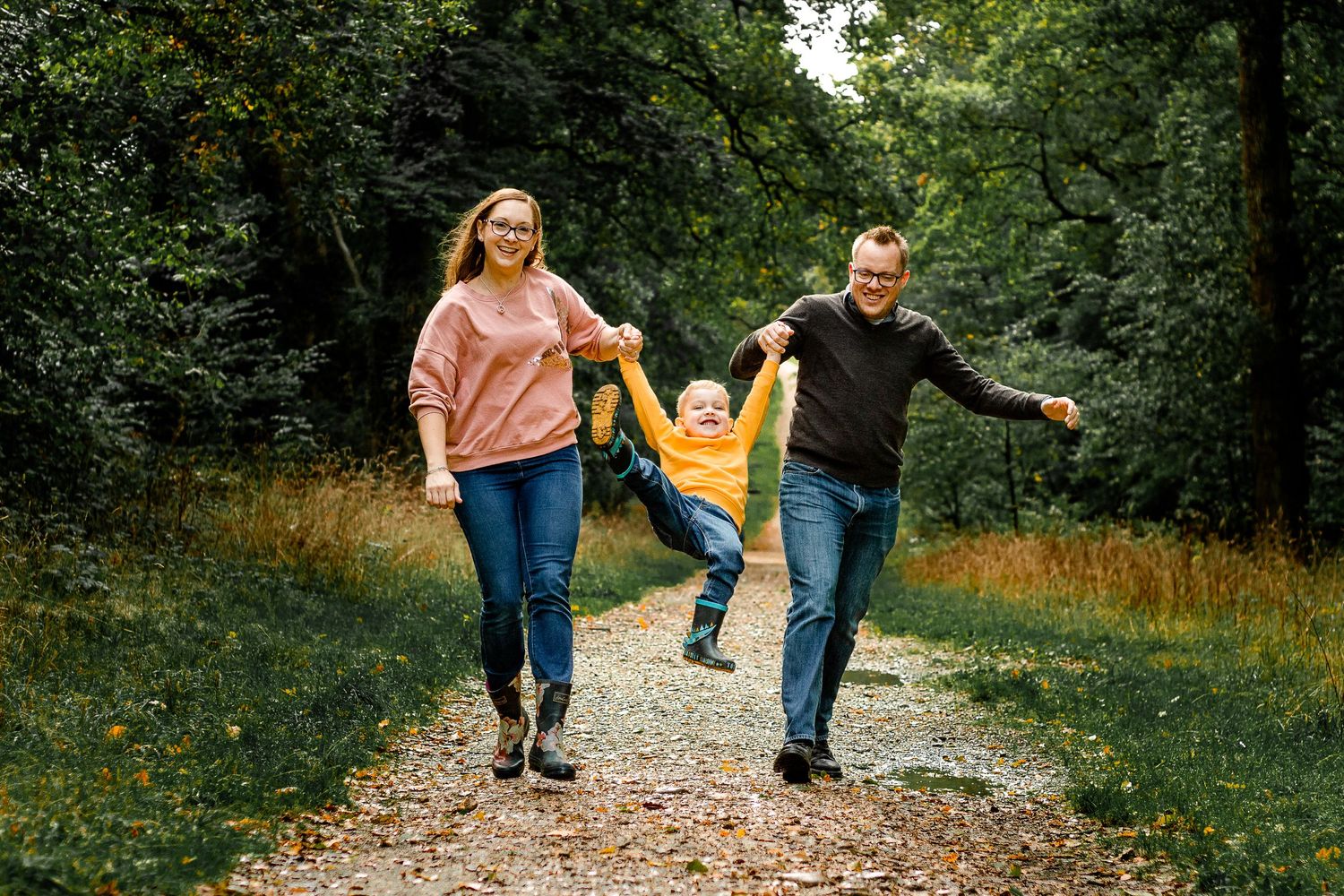 In the woods at Staunton Country Park, Hampshire, parents swing their 5 year old between them laughing.