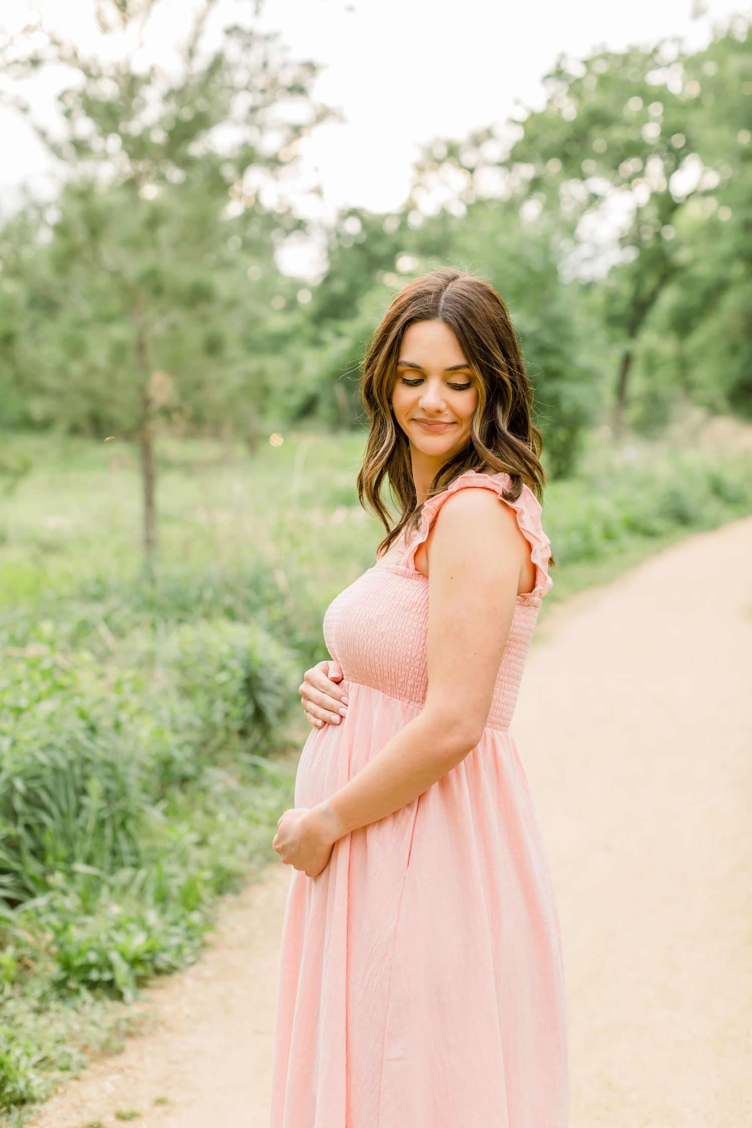 A woman in a flowing pink dress cradles her baby bump on a scenic nature path.