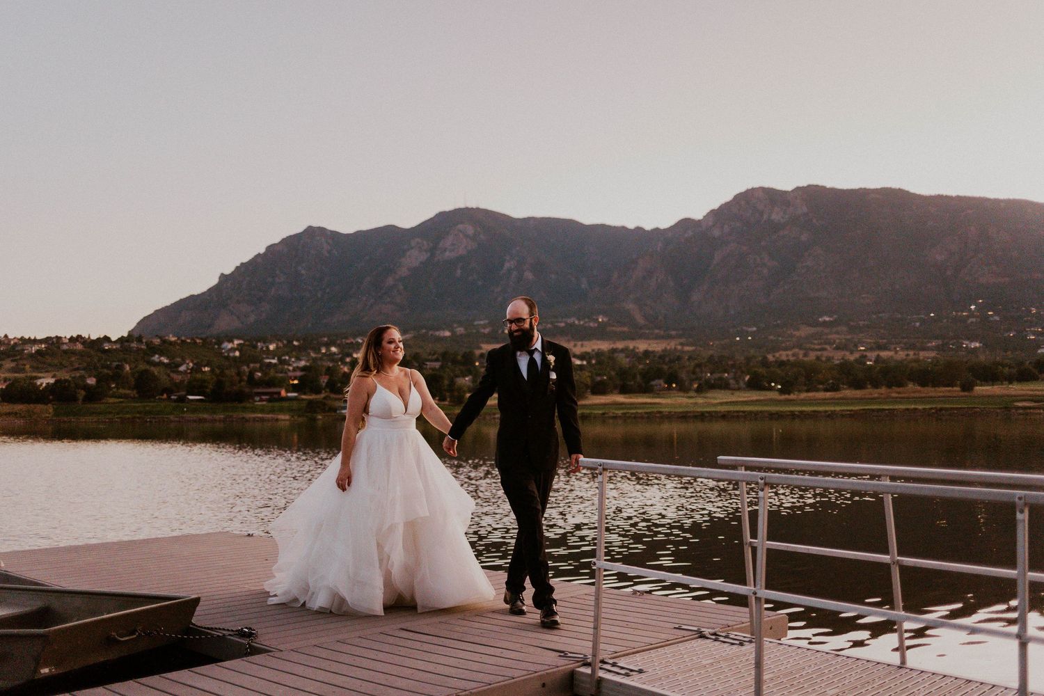 A couple walks along a lakeside dock with mountains in the background at sunset.