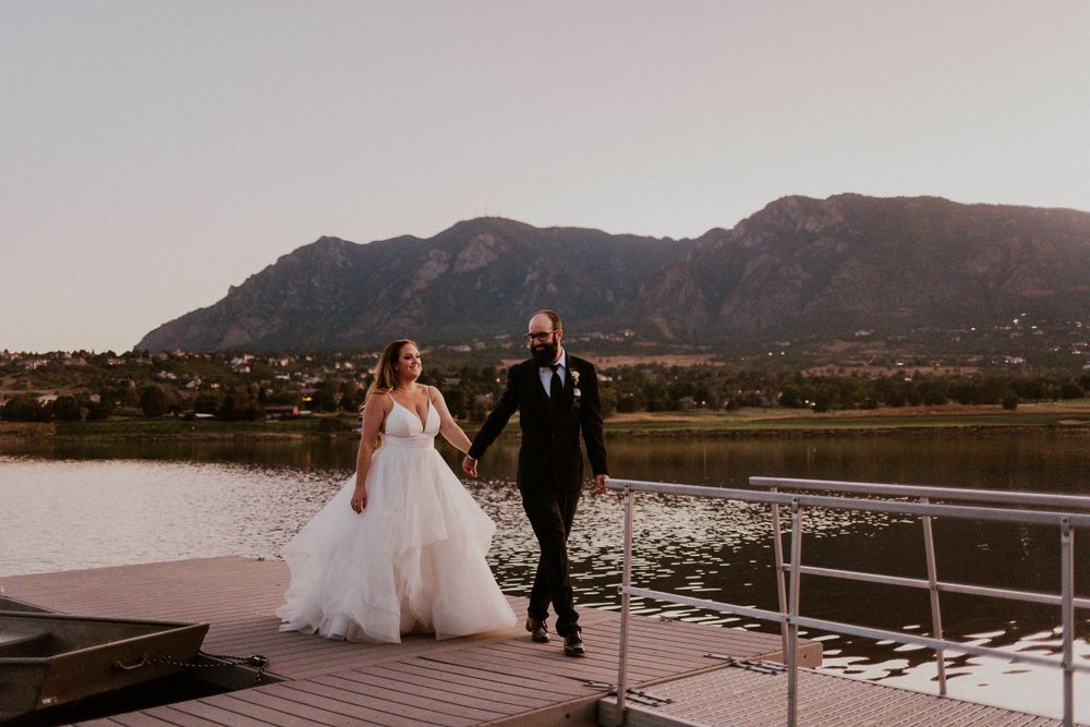 A couple walks along a lakeside dock with mountains in the background at sunset.